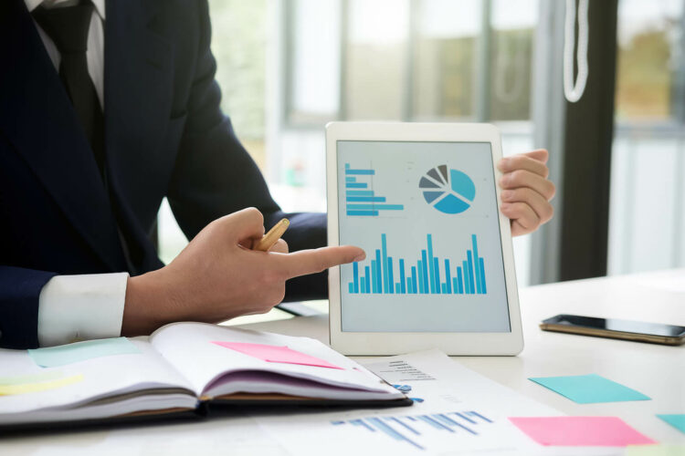 Businessman sitting at desk with papers scattered and pointing at iPad displaying charts.