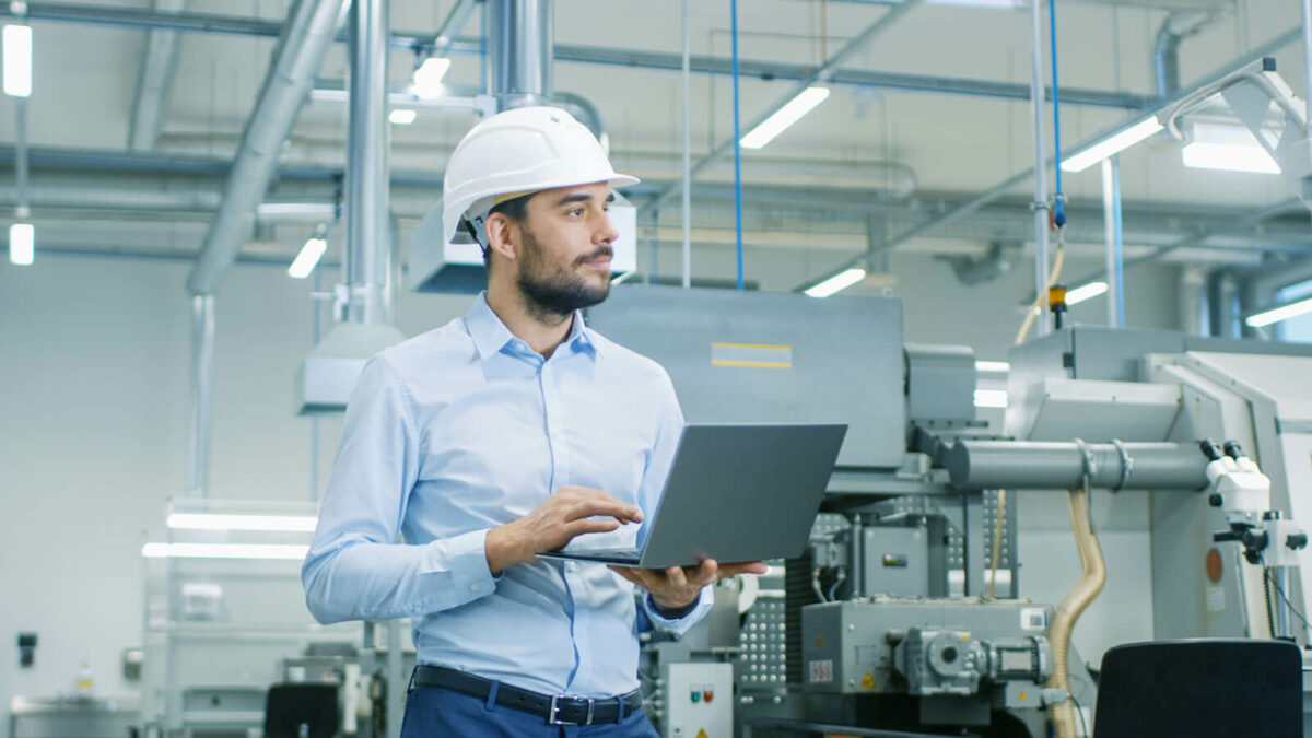 Man with hard hat on is holding his laptop in manufacturing plant or factory.