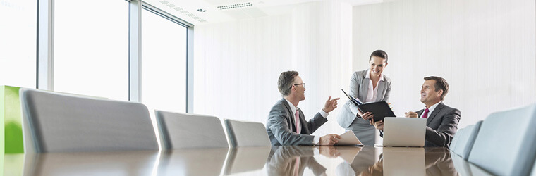 Three individuals sitting at the end of a conference table discussing papers in folder.